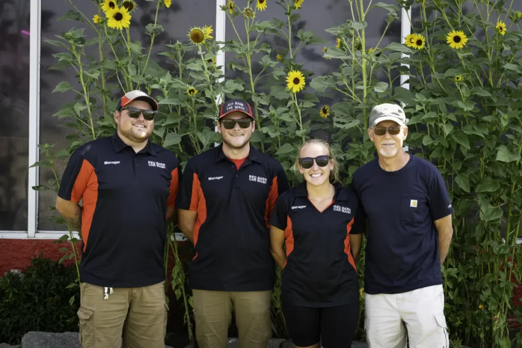 red-barn-car-wash-287-team red barn car wash staff in front of yellow flowers in front of the tunnel car wash building